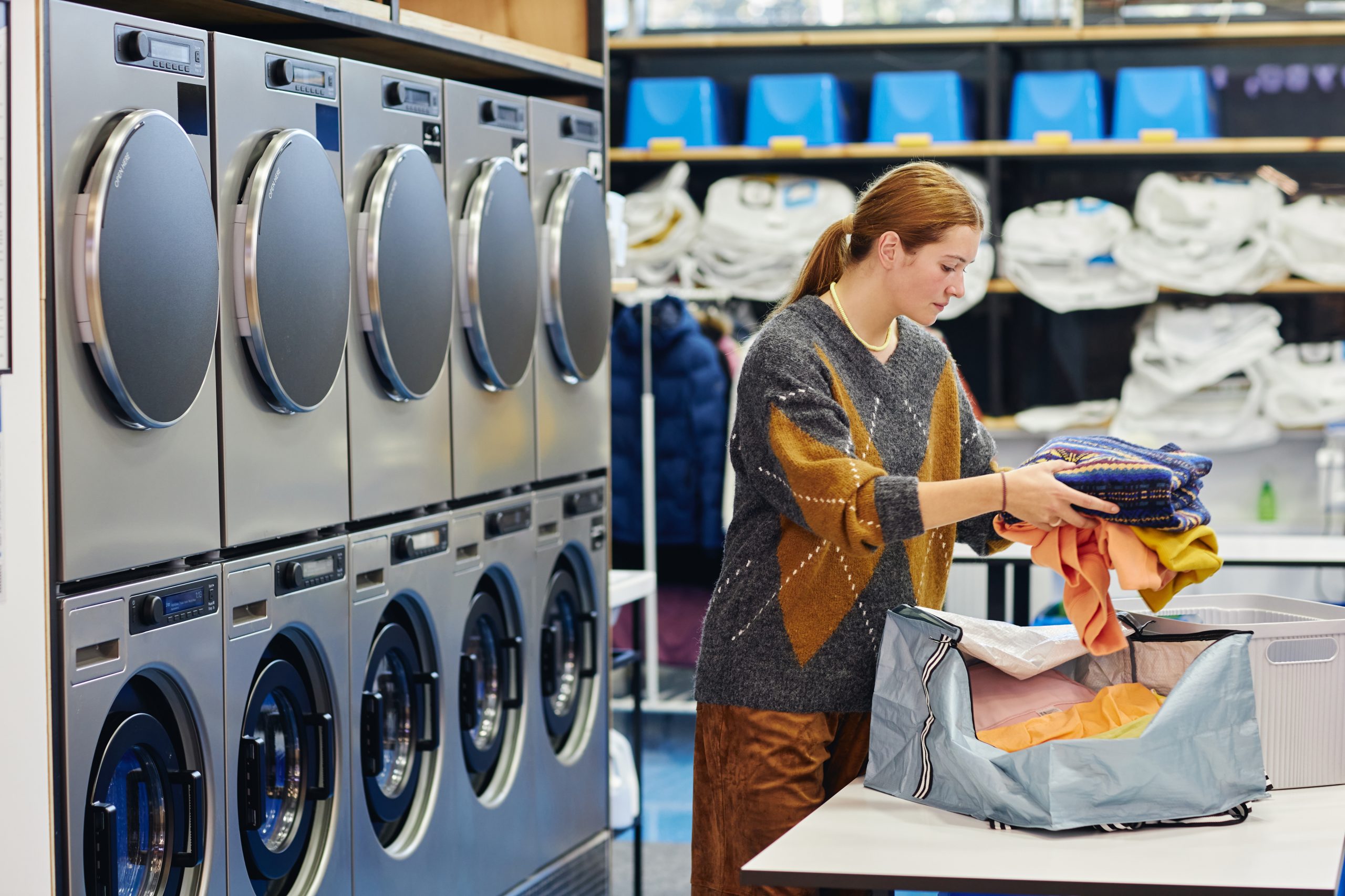 Side view of woman putting pile of sweaters into basket on folding table while preparing to load clothes in washing machine in self service laundry room, copy space